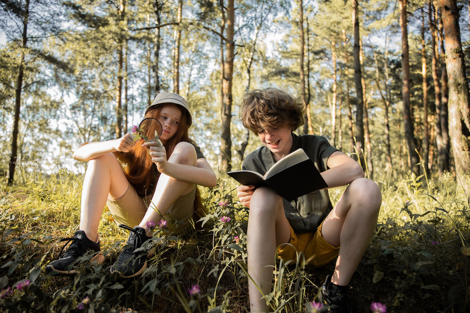 Pikkade punaste juustega tüdruk ja lokkis juustega poiss istuvad matkariietes metsas maas ja uurivad loodust. Noorte teadlaste klubi